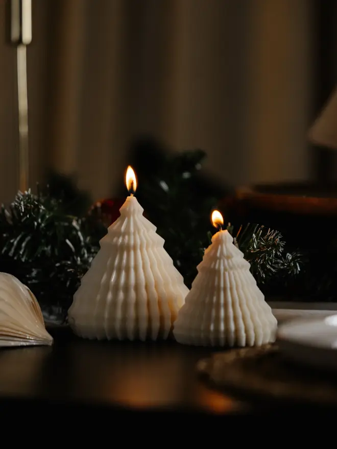 Origami Christmas Tree Candles (Set Of 2). Two lit cream cone tree candles with intricate fluted ridges, glowing beside frosted pine garland and a paper ornament on a dark table. The textured sculptural design gives a soft, cosy glow to winter evenings.