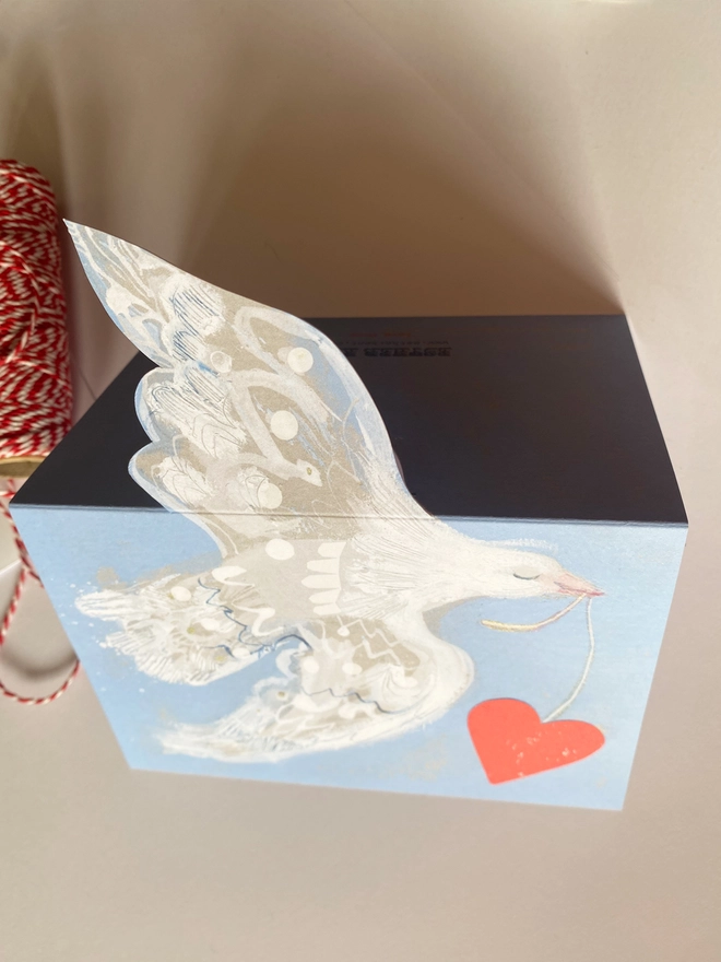 Esther Kent greetings card showing white flying dove in a light blue background, carrying a vibrant red heart on a string