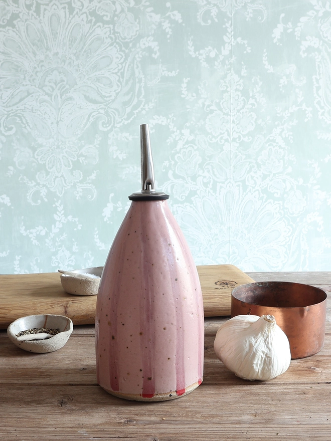 Handmade Ceramic Stoneware Oil Pourer in Pink Stripe sat on a wooden surface next to a bulb of garlic, a brass measuring cup, a wooden chopping board and small ceramic dishes containing salt and pepper. 