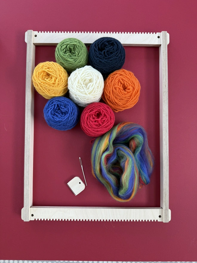 Wooden beige frame loom with seven balls of yarn in white, green, blue, orange, yellow, red and cream plus some rainbow merino fibres, awooden comb and needle