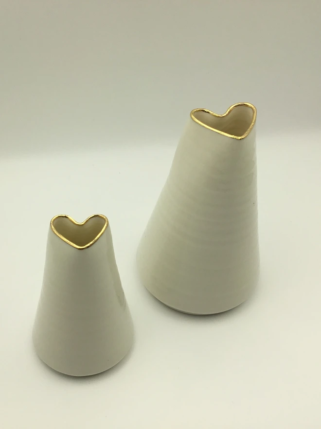 porcelain heart shaped bud vase. two white porcelain bud vases photographed on a white backdrop with a gold rim.