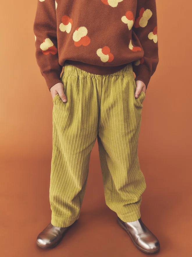 Child stands facing camera against a burnt orange backdrop, wearing a Studio Koter chestnut knitted jumper with yellow and orange floral intarsia and chartreuse wide-leg corduroy trousers.