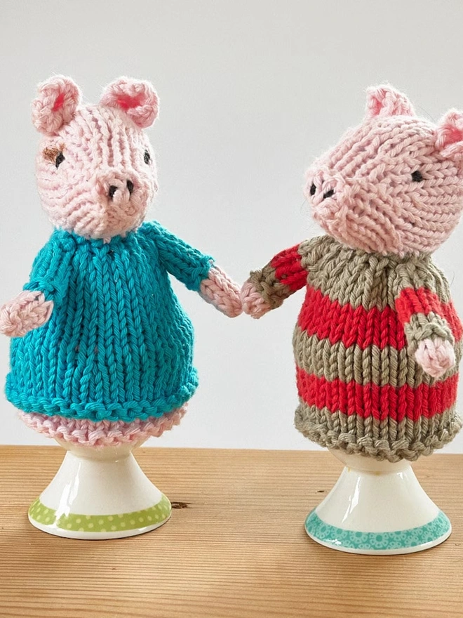 Egg Cosy Pigs Table Decoration (Set Of 2), a pair of colourful pig cosies sitting on a wooden surface. 