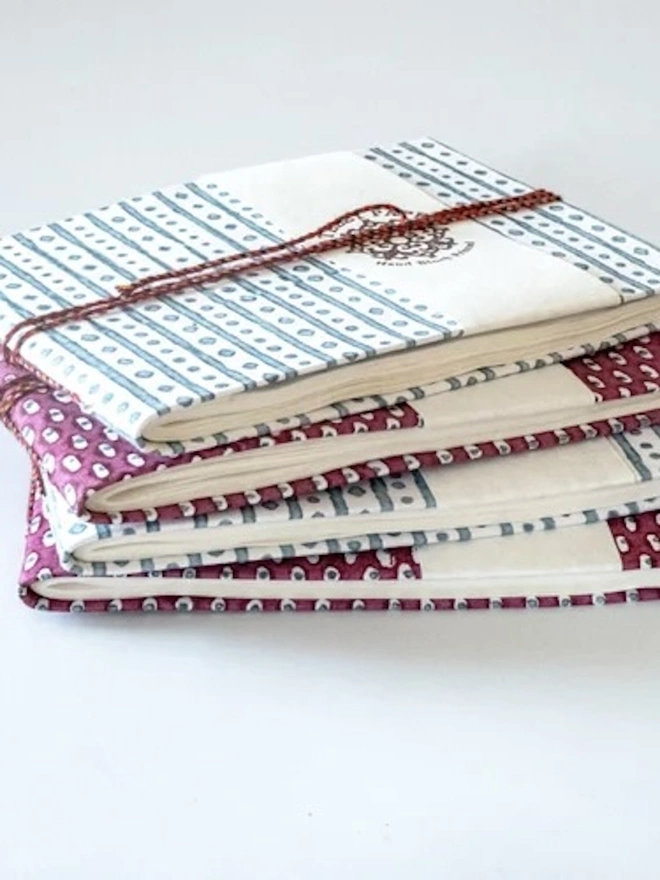A5 Small Landscape Travel Watercolour Book. A stack of hand block printed notebooks in slate blue stripe and burgundy spot designs, tied with red braided cord. The handmade journals have cream paper pages and a Yateley Papers branded band for a thoughtful stationery gift.