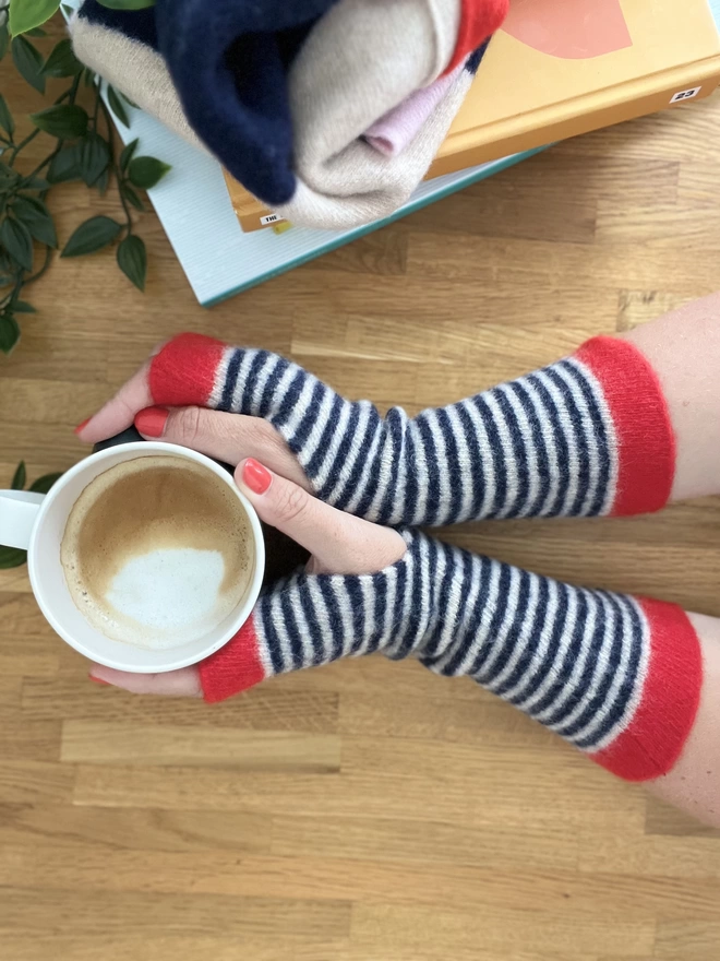 Knitted wristwarmers in red and navy being shown worn and holding a coffee cup