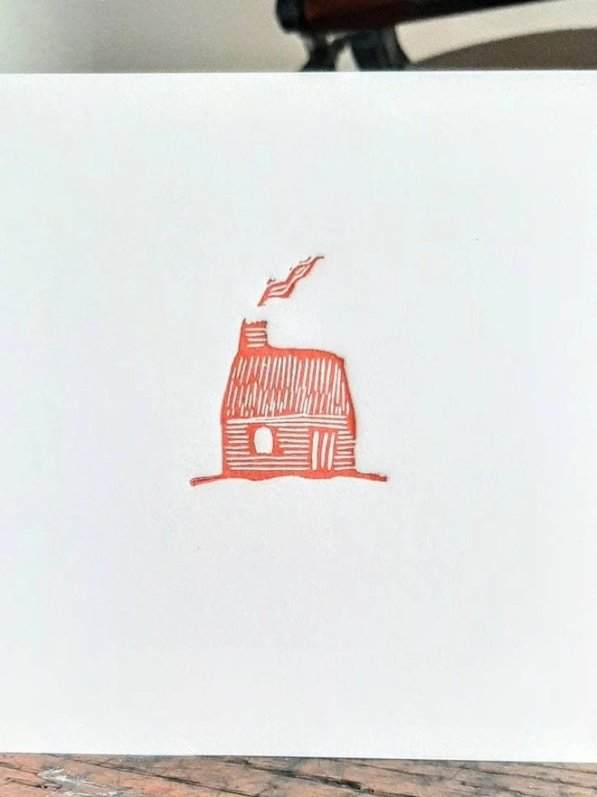 Tiny Red Cottage Letterpress Handprinted Greetings Card, a white card with a tiny red cottage design in the centre, on a wooden surface. 