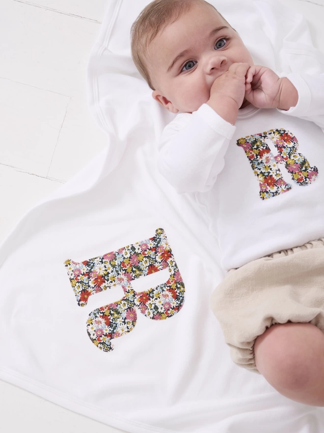 A baby laying on a soft white cotton blanket with an appliqué Liberty print initial on it