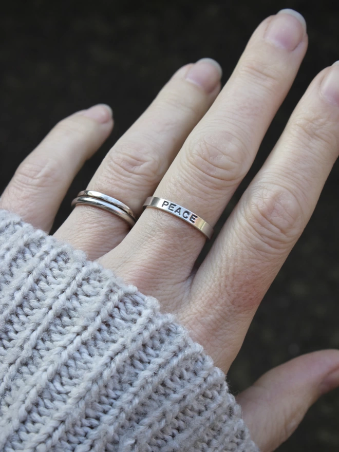A sterling silver ring band with the word 'peace' stamped on it, worn on the left hand of a woman wearing a grey knitted jumper.
