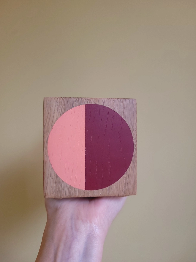 Wood cube tealight holder painted with a red and pink circle O helod against a yellow wall