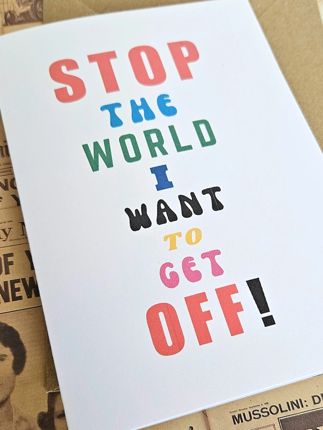 A mindful empathetic greeting card that says stop the world I want to get off in colourful fonts on a white card photgraphed on a background of old newspapers
