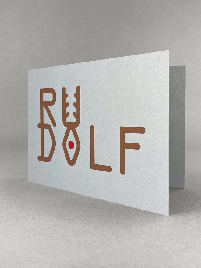 Rudolf is spelt out in brown ink, stacked to look like a reindeer - with a red dot depicting the nose. The grey card is stood in a light grey studio set, slightly open.