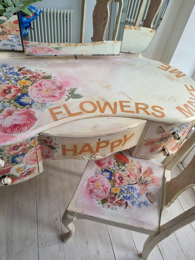 An antique kidney-shaped dressing table with chair and a trio of mirrors. Upcycled in cream and pale pink with decoupage florals in oranges and pinks. Text across the table read ‘flowers make me happy’ in the orange of the undercoat.