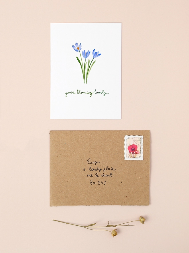 An image of a mini, rectangular white greetings card with an illustration of three blue crocus flowers with a hand written 'you're blooming lovely' message seen under the central illustration. The mini greetings card is placed on top of a rectangular brown kraft envelope.