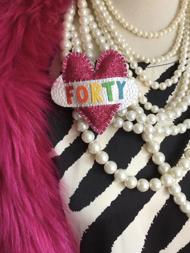 Pink glittery heart shaped brooch with FORTY spelt out in rainbow coloured lettering on a white scroll, pinned to a zebra dress, with pearl necklace and hot pink fur stole