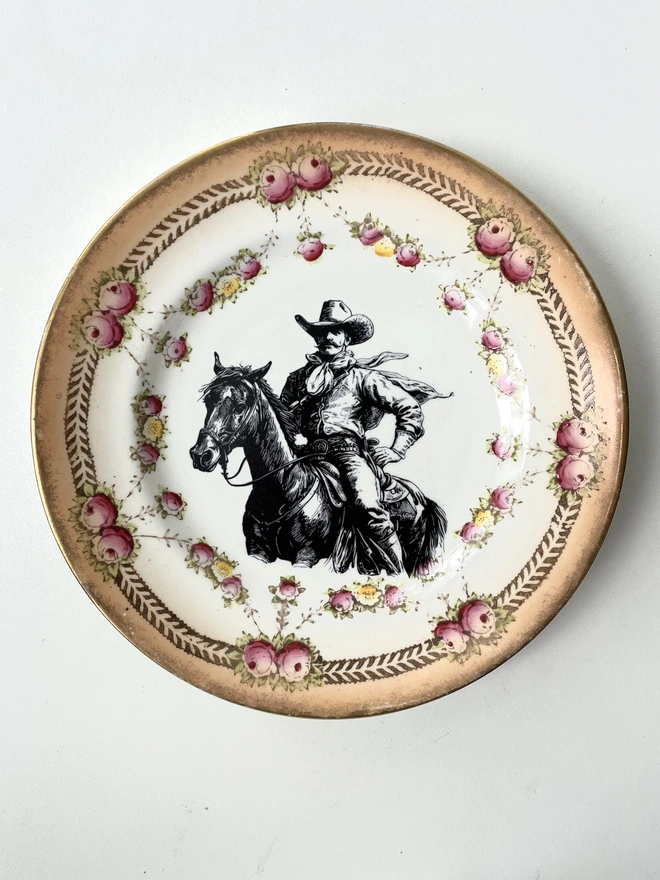 vintage plate hanging on a white wall, it has a decorative gold edge and floral border, with a printed vintage illustration of a cowboy in the middle 