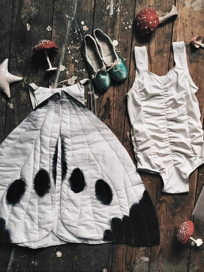 Cabbage white butterfly wings costume placed flat on wooden boards with a white ruched leotard, mint green ballet slippers, and scattered red and white toadstools