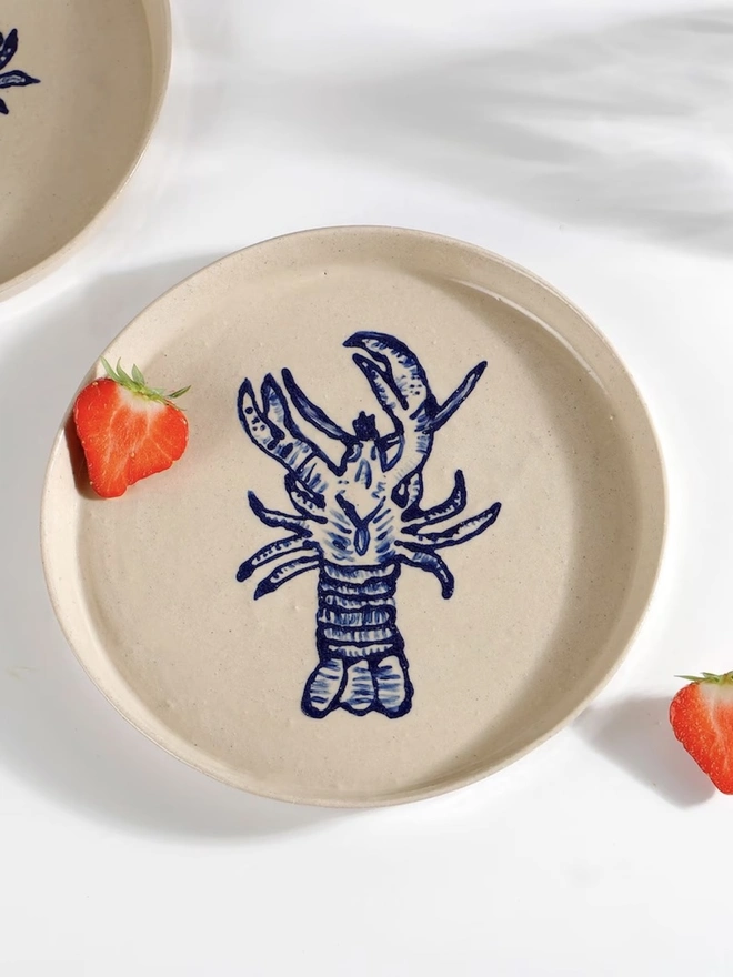 Handmade Ceramic Side Plate With Blue Lobster Design, a side plate with a blue lobster design sitting on a white table surface accompanied by a raspberry. 