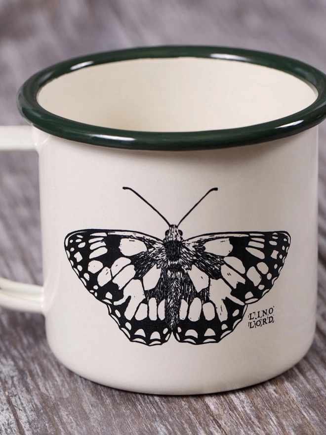 Picture of a Cream Enamel Mug with a Green Rim with a butterfly design etched onto it, taken from an original Lino Print