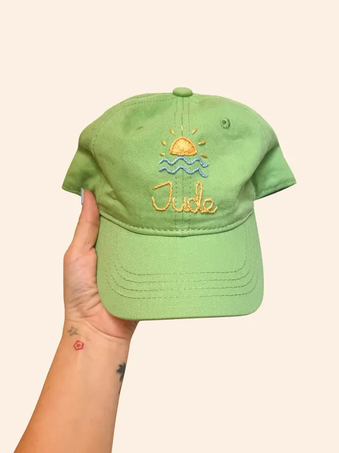 Hand Embroidered Kids Cap, a person holding a personalised cap against a colourful backdrop. It says a child's name. 