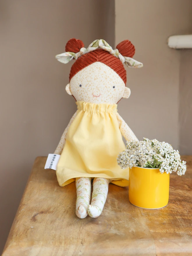 Handmade heirloom cloth doll with freckled face, red corduroy hair in two buns, floral leggings, and a soft yellow dress, styled on a wooden table with delicate wildflowers.