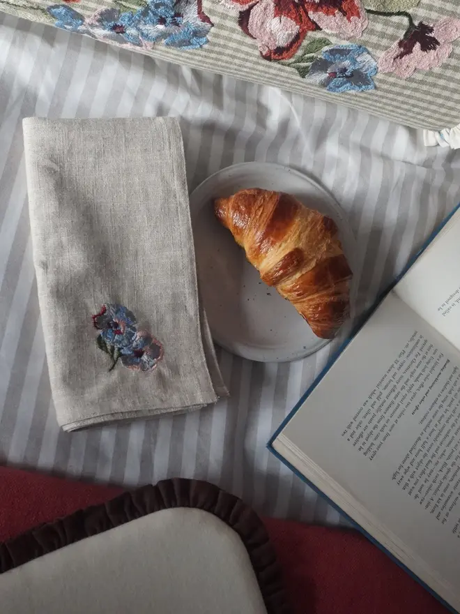 Natural linen napkin embroidered with blue and pink flowers, styled for breakfast with coordinating embroidered homeware.