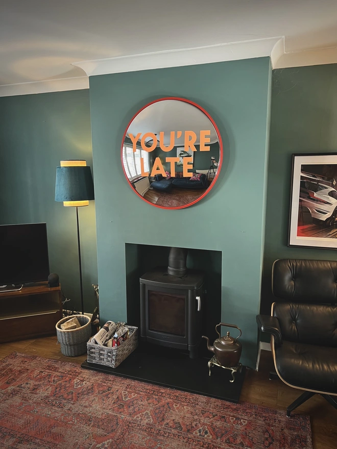 Round convex mirror with 'You're Late' hand painted in orange in a red round frame, on a teal wall and log burner, with a lamp, logs and white ceiling