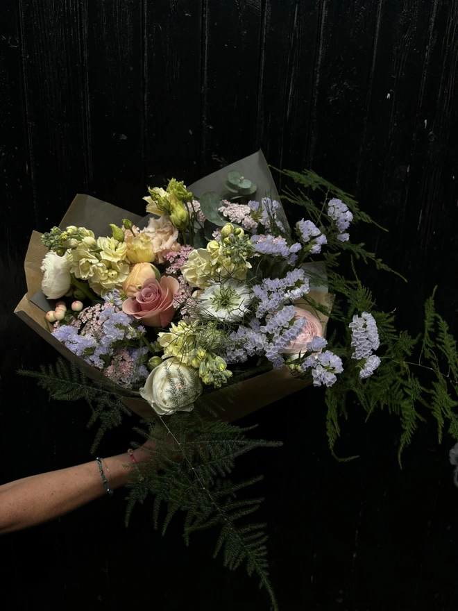 A bouquet of pastel hand tied flowers being held up on a dark background with gift wrap