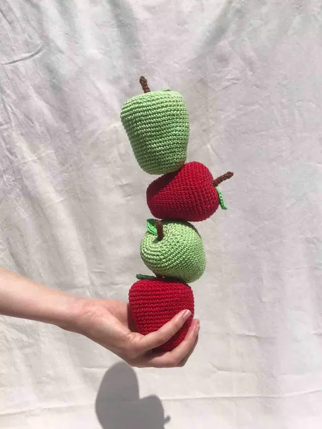 Image of two green and two red crochet apples balanced on top of each other and balanced on a hand with a plain background.