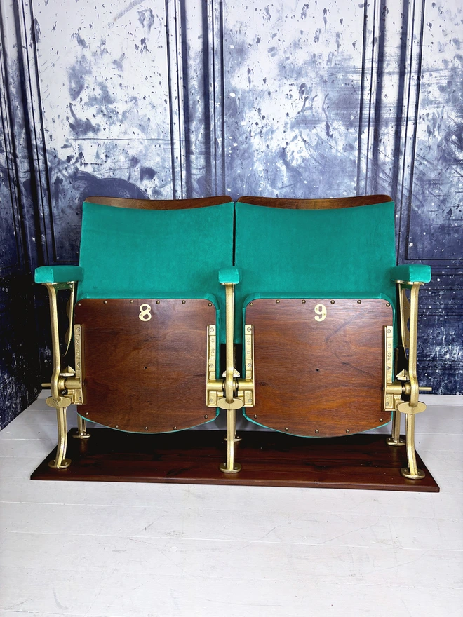 Set of two vintage refurbished cinema seat upholstered in a beautiful green plain velvet.  The cinema seats have gold legs and are mounted onto a wood plinth.  The flip down part of the seats are closed showing their wooden bases.