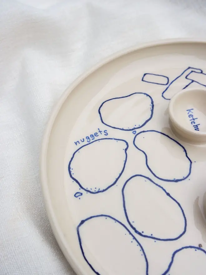 Dippy Chips & Nuggets Dinner Plate, a white dip dinner plate with blue outlines for nuggets and chips sitting on a white bed cloth. 