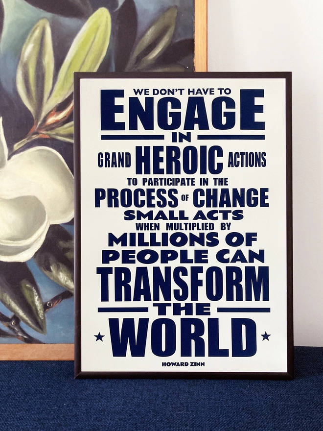 Framed typographic print, with navy blue text on a white background. Quote by Howard Zinn - “We don't have to engage in grand, heroic actions to participate in change. Small acts, when multiplied by millions of people, can transform the world.” The print rests on a painting of white flowers on a dark blue background.