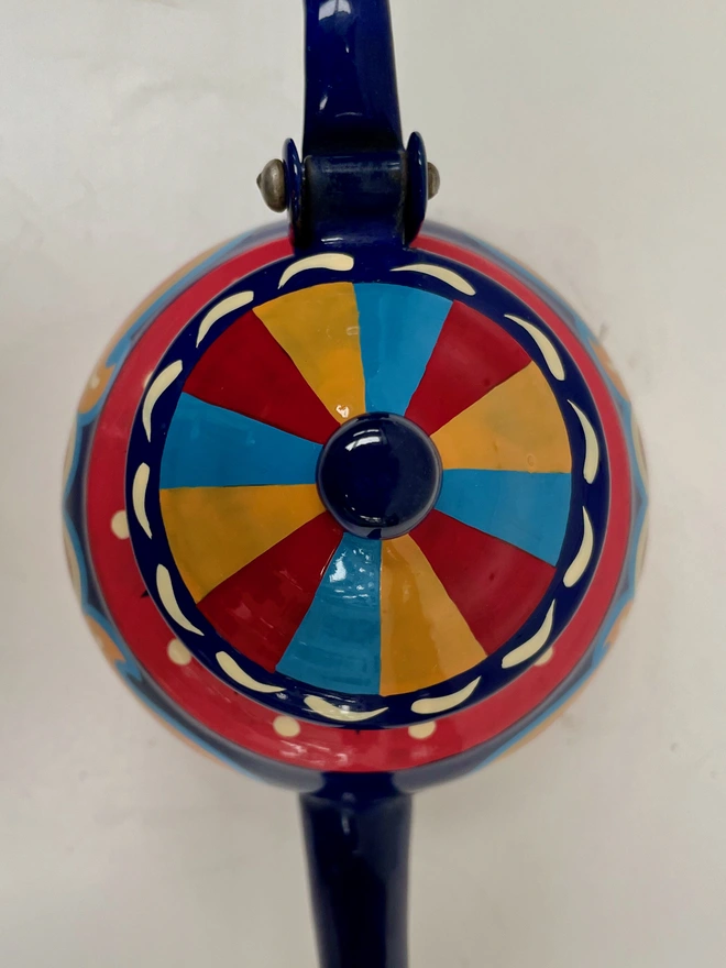 Lid of small blue teapot, featuring a yellow, red and blue striped design.