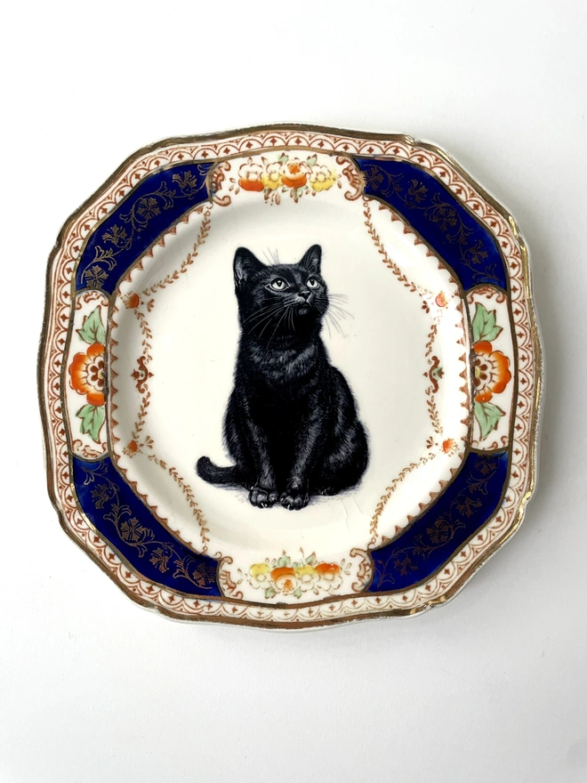 a vintage plate with an ornate border, in the centre of the plate is a vintage style black and white illustration of a black witch's cat