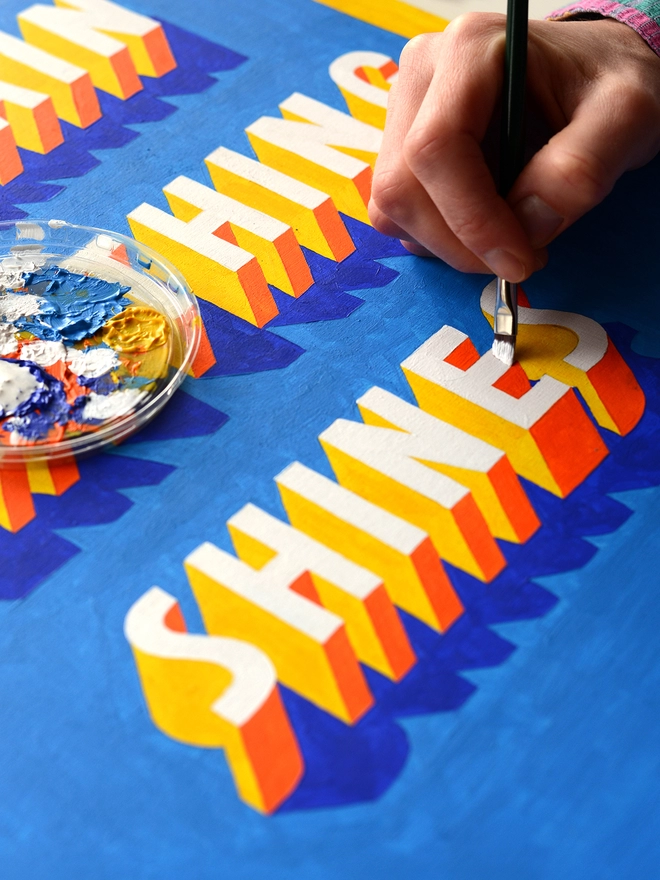 Close up of an artist's hand painting 3d typography of the word Shines