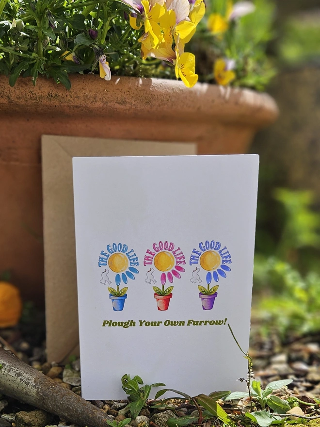 A white card with an illustration of  3 colourful flowers each in a terracotta pot displayed in a row with a white bird. The Good Life is written using a retro style font in 3 colours. It says at the bottom of the card Plough Your Own Furrow!