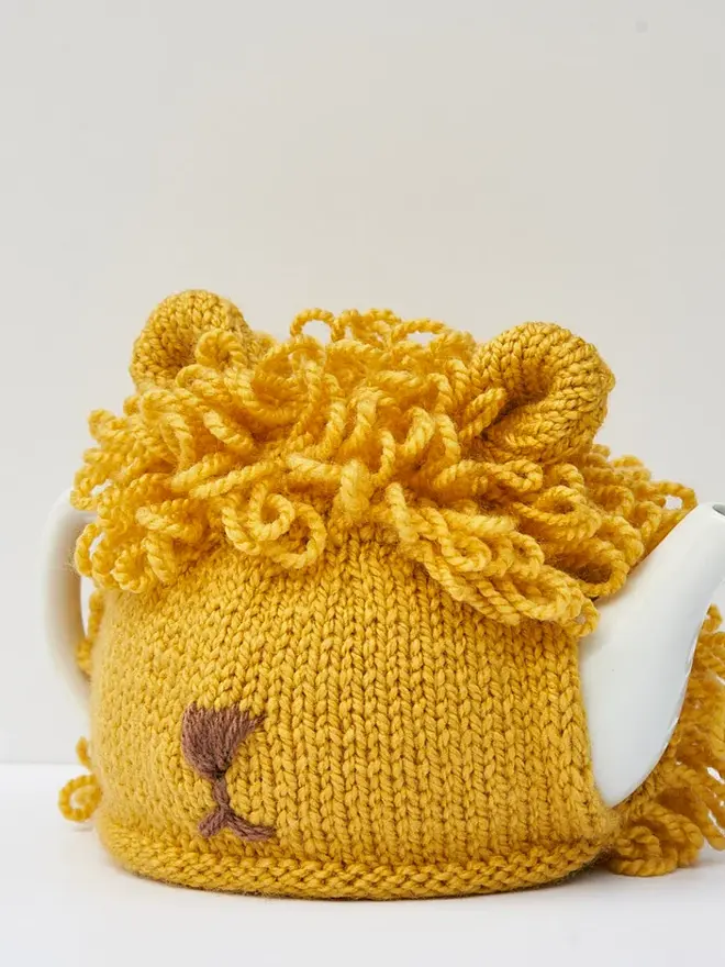 Lion Tea Pot Cosy Animal Lovers Tea Time Treat, a yellow lion tea pot cosy sitting on a tea pot, on a white surface against a grey backdrop. 