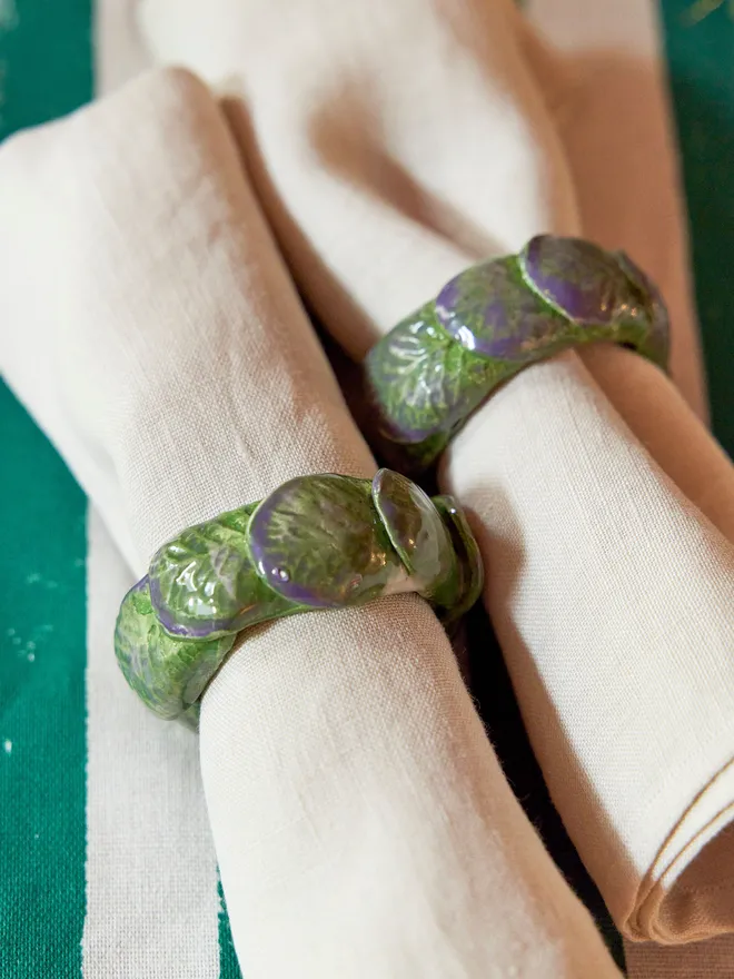 Close-up of two handmade ceramic sprout napkin rings around natural linen napkins, showing their glossy overlapping leaves in mottled green and purple glazes.