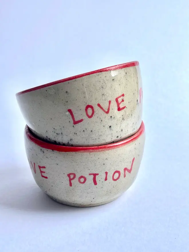 'Love Potion' Mug, two ceramic mugs with 'Love Potion' written on them, stacked on top of each other in front of a blue background. 