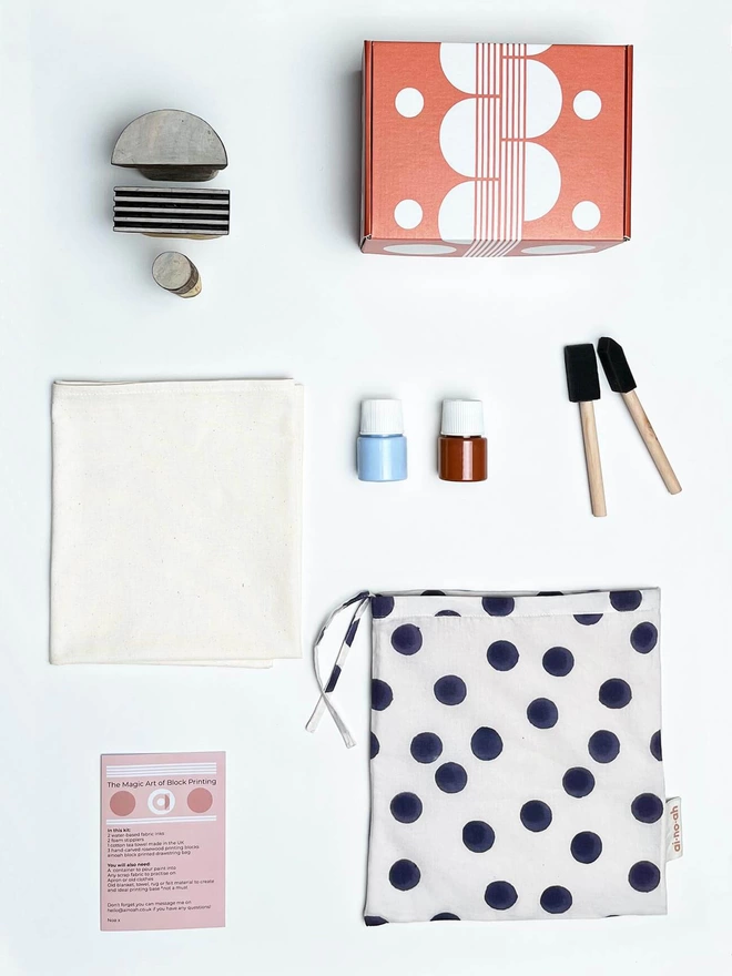Block printed bag, a pot of blue ink and a cinnamon coloured one, two foam stipplers, a plain cream tea towel, three wooden blocks for printing and a soft orange printed cardboard box on a white surface 