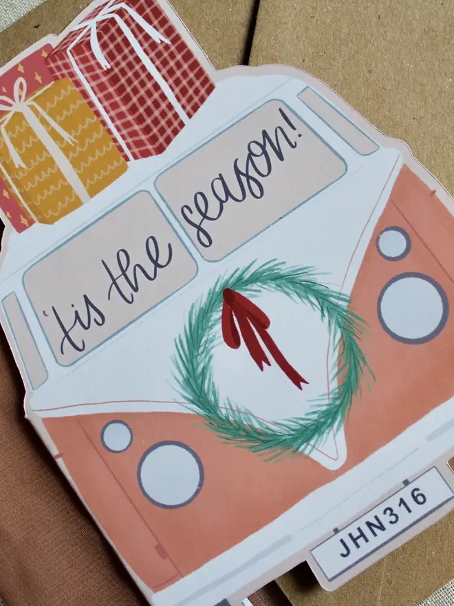 tis the season illustrated campervan christmas card. pink illustrated campervan card emblazoned with phrase tis the season and decorated with christmas gifts and a wreath.