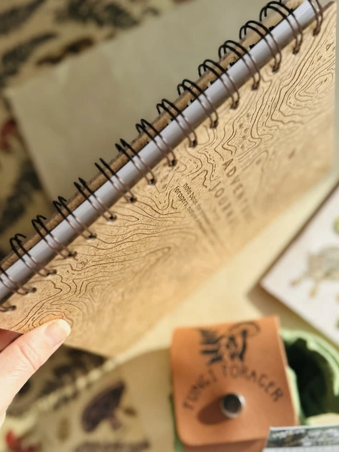Wooden Hardback Adventure Journal, a person holding an adventure journal to its side against a colourful backdrop. 