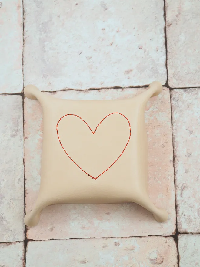 The base of a heart leather tray. The underside of the tray shows the square shape and natural cream colour. A red stitch line in the outline of a love heart shape shows how the interior heart is hand stitched to the tray.