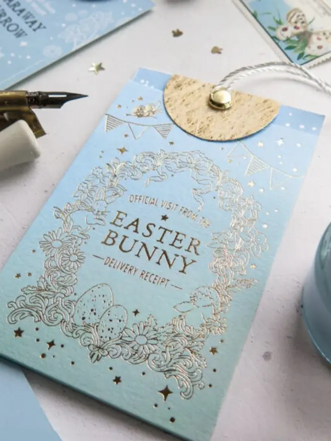 Close-up of the gold foil "Easter Bunny Delivery Receipt" lettering and bunting on the blue pouch. A brass dip pen nib rests beside it.