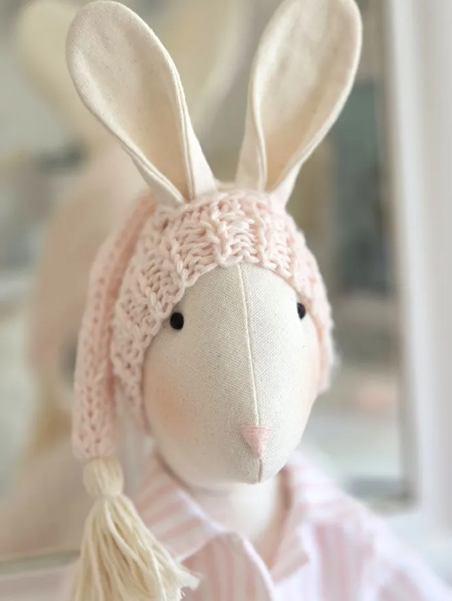 Close-up of handmade linen Veronica Bunny wearing a pale pink hand-knitted sleepy hat with cotton tassel and pink striped pyjamas.