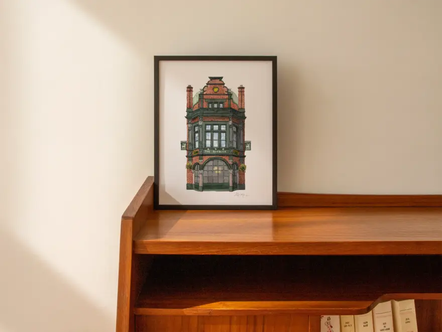 Borough The Roebuck Pub Giclée Print (Unframed), a print of a pub sitting in a black frame on a wooden surface against a plain backdrop.