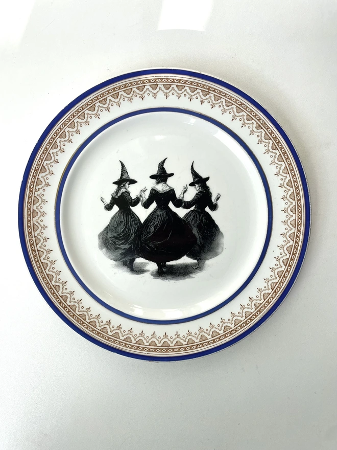 a coven of 3 witches dance hand in hand, the image is victorian in black and white and is printed directly onto an ornate vintage plate