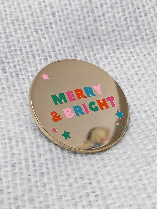 A gold enamel pin with pastel lettering that reads "Merry & Bright" lays on a knitted fabric blanket.