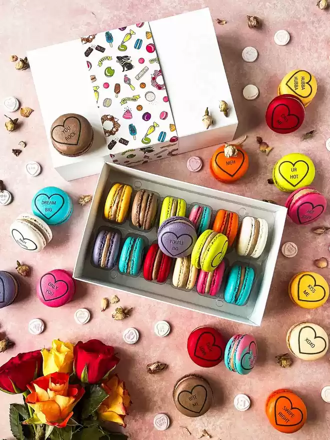 Colourful macarons decorated like conversation love heart candy in a white gift box on a pink background with colourful roses