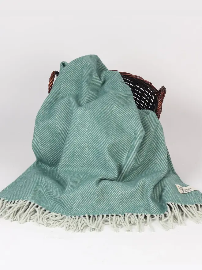 Pistachio Recycled Wool Tartan Blanket, a blue picnic blanket sitting on a white surface against a wooden basket, against a plain backdrop. 