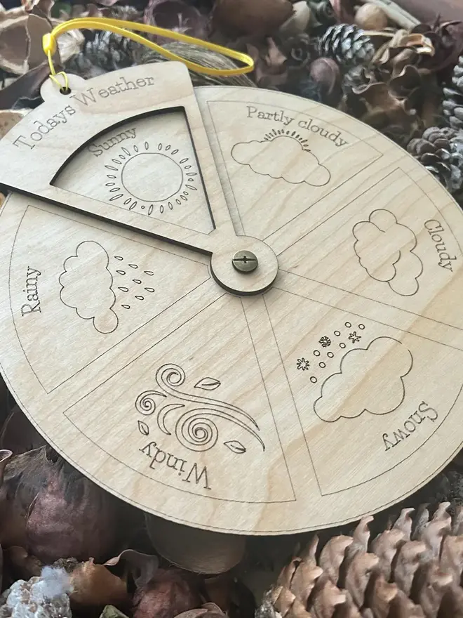 Extra large wooden weather wheel with engraved weather symbols for sunny, cloudy, rainy, windy, snowy, and partly cloudy, shown resting among pinecones and natural materials.